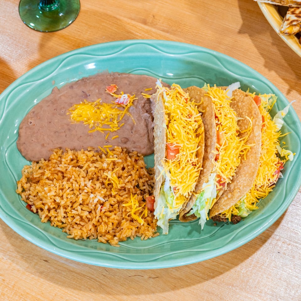 Three crispy tacos with lettuce, cheese, and tomatoes, served with refried beans and rice.