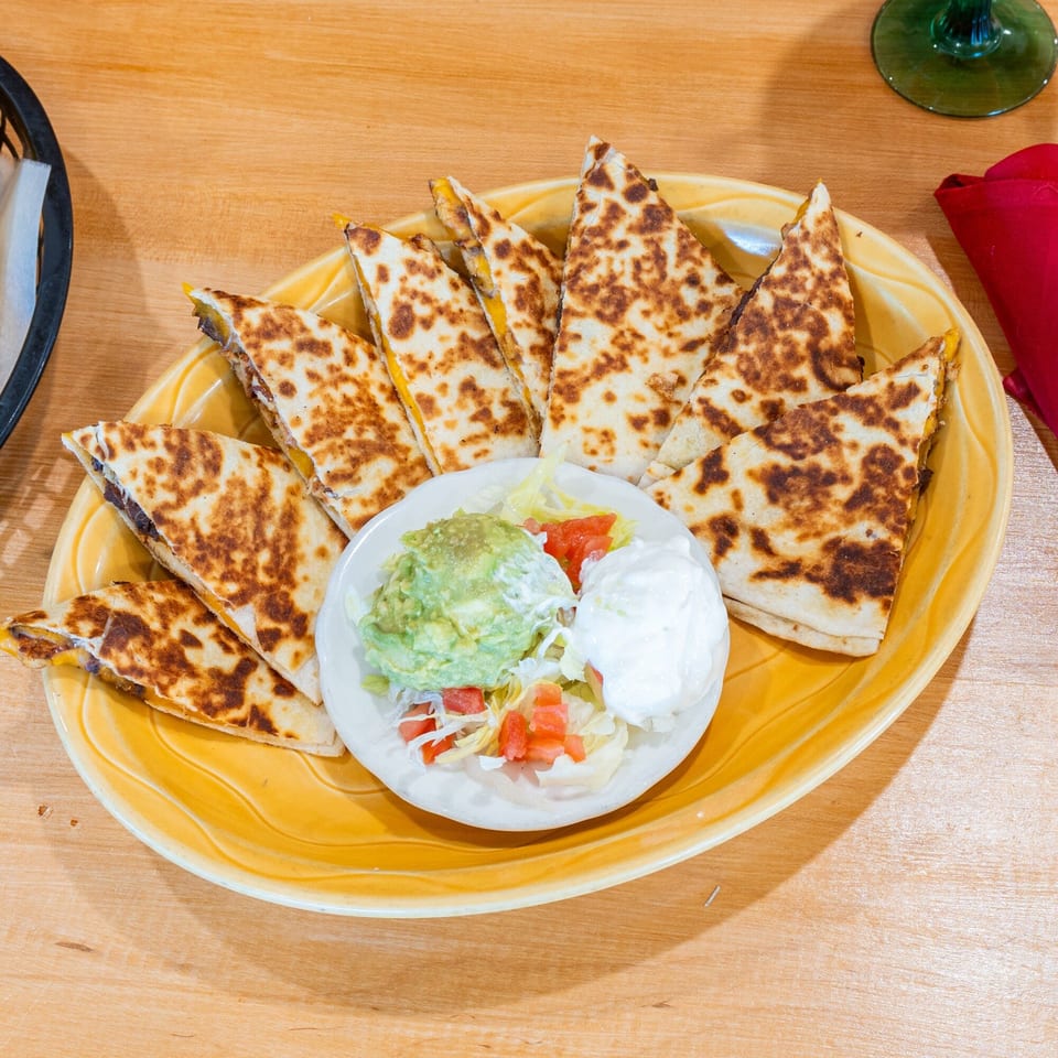 A yellow plate of quesadilla wedges with a side of guacamole, sour cream, and diced tomatoes.