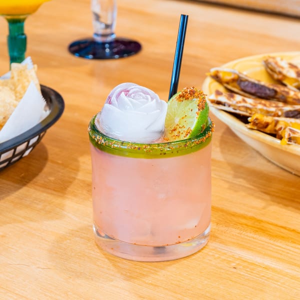 Pink cocktail with a white rose ice cube, lime, and chili salt rim on a wooden table.