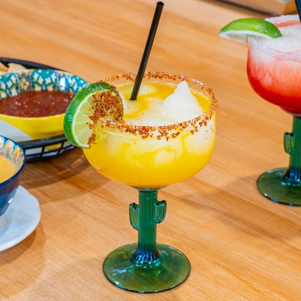 Two colorful frozen margaritas in cactus-stemmed glasses, with salsa and chips in the background.