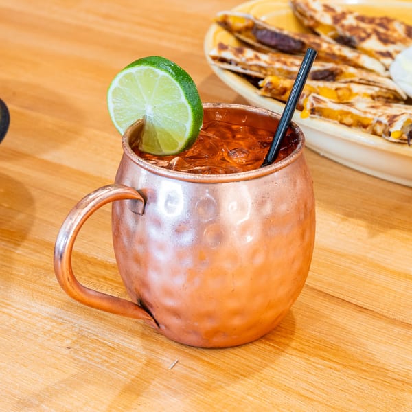 A copper mug with a drink, lime, and straw, next to quesadillas on a wooden table.