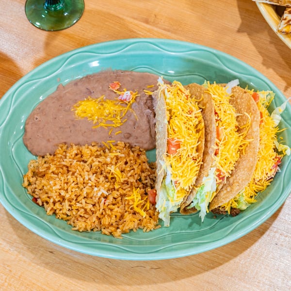 Three crispy tacos with shredded cheese, lettuce, and tomatoes, served with rice and refried beans.